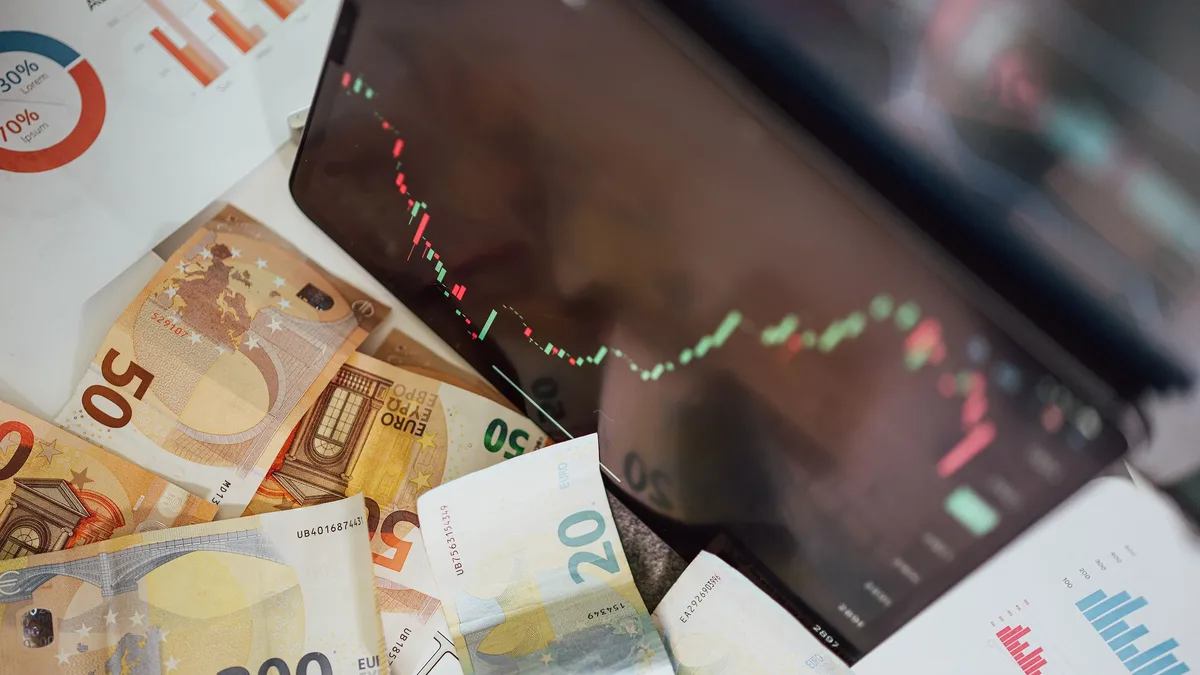 A collection of Euro banknotes and a blurred stock market graph on a computer screen.